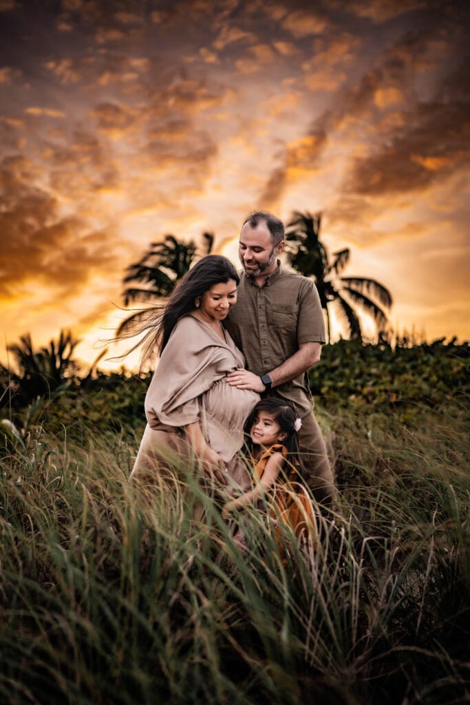 Maternity Photographer, a man, wife and daughter, hold each other in tall grass in a tropical area. The woman is pregnant