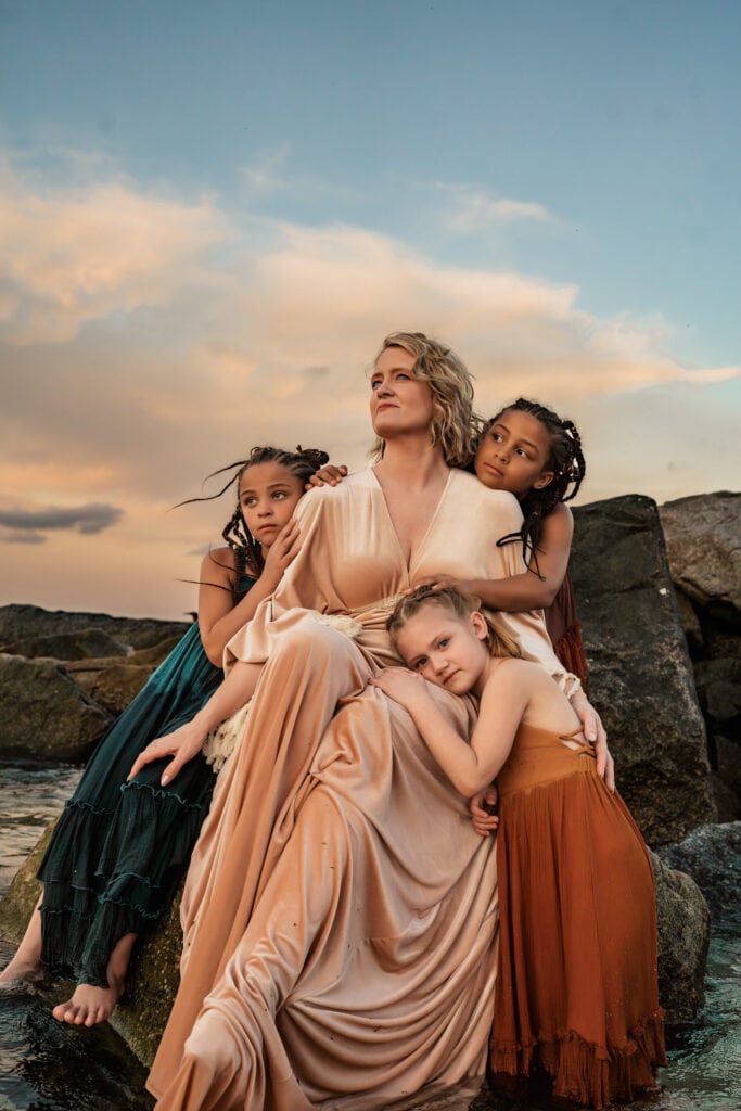 Family Photographer, 3 girls hug their mother, they all wear dresses and lean on beach rocks